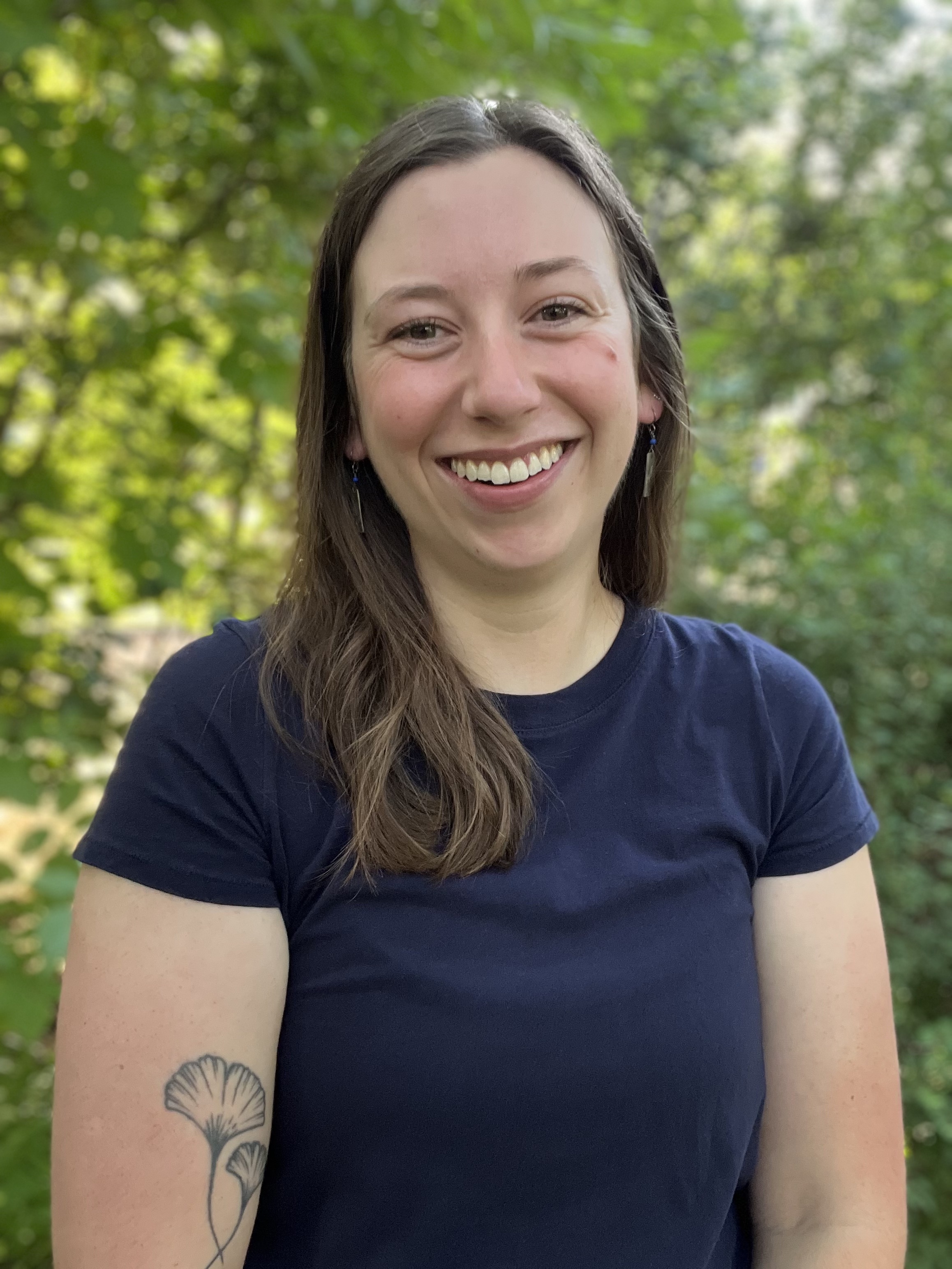 Photograph of Aly Ennis from the torso up in front of foliage.