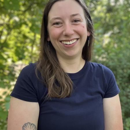 Photograph of Aly Ennis from the torso up in front of foliage.