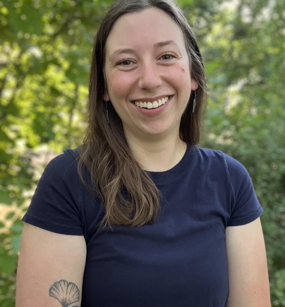Photograph of Aly Ennis from the torso up in front of foliage.